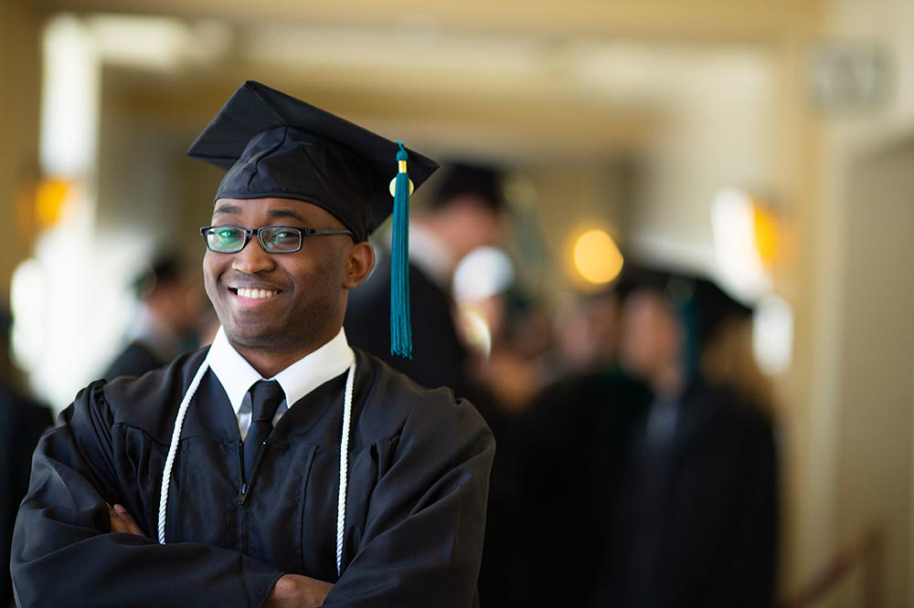 Boy with arms folded, smiling wearing his graduation cap and gown.