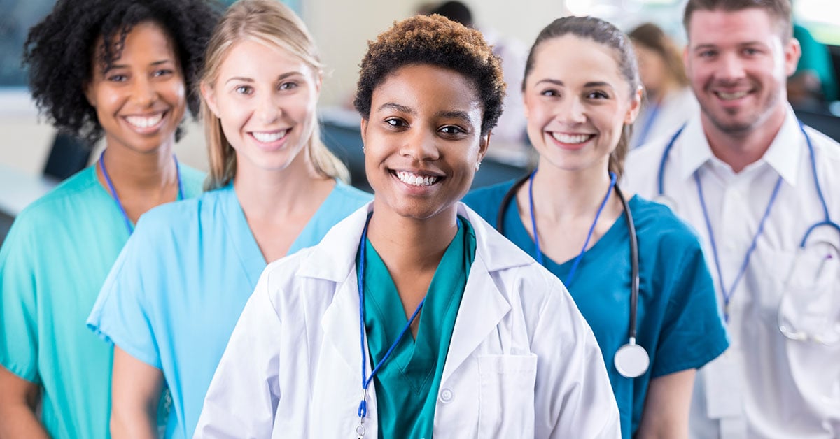 Five nurses, four female and one male, in a V-shape are facing forward and smiling.