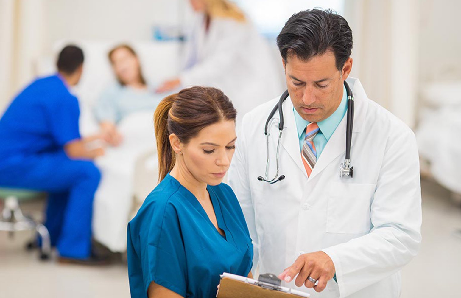 Female in blue scrubs looks at clipboard with male doctor standing next to her