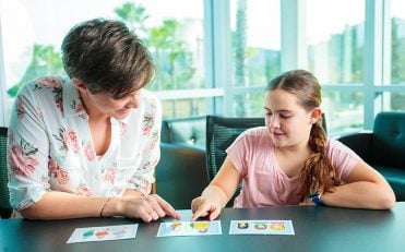 Female speech therapist seated with young girl at table pointing at flashcards