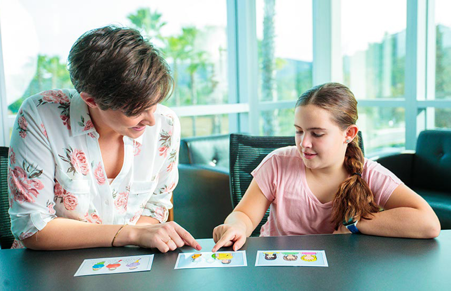 Female speech therapist seated with young girl at table pointing at flashcards