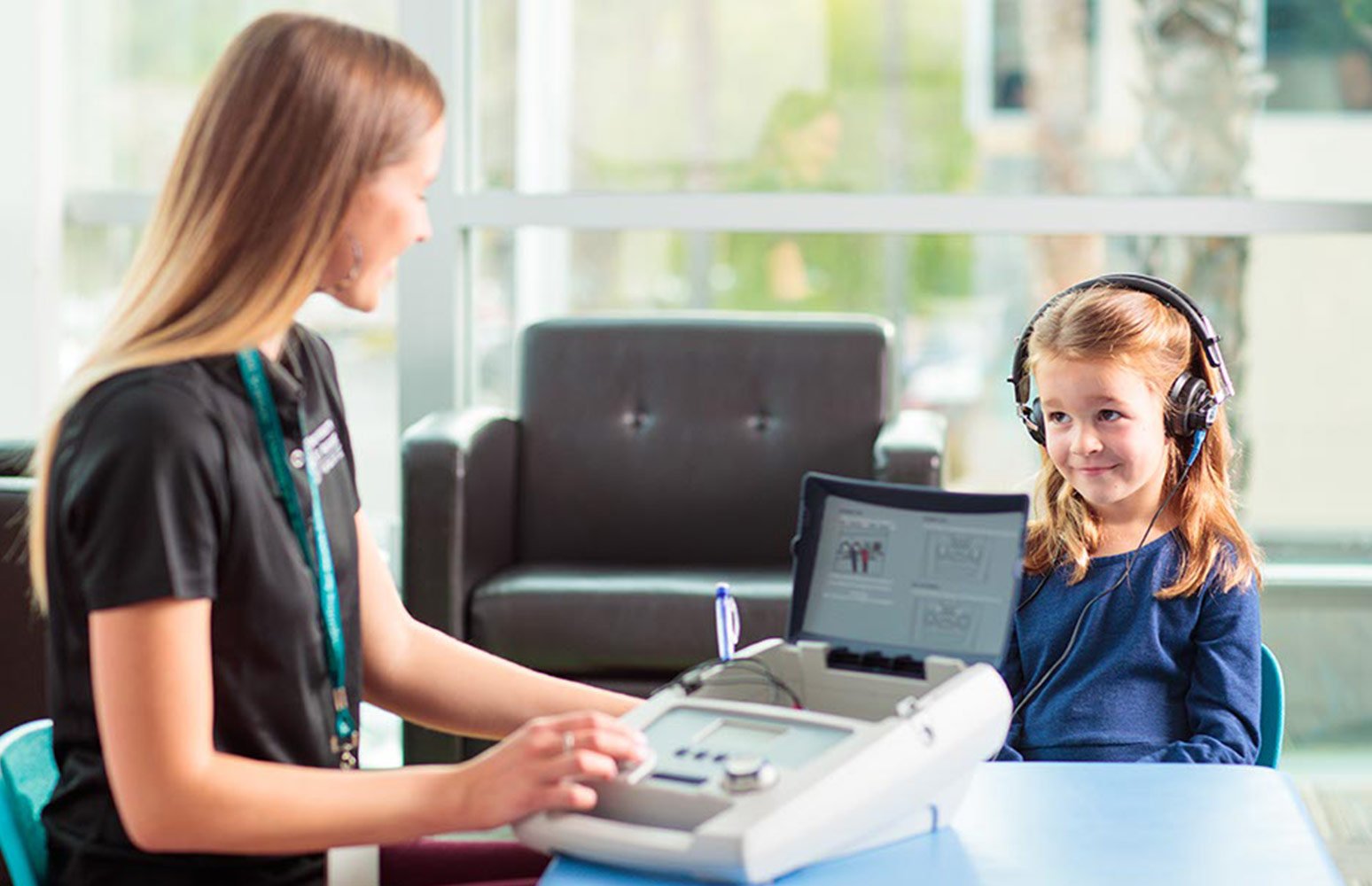 Girl with headphones on looks at woman turning knob on auditory testing machine